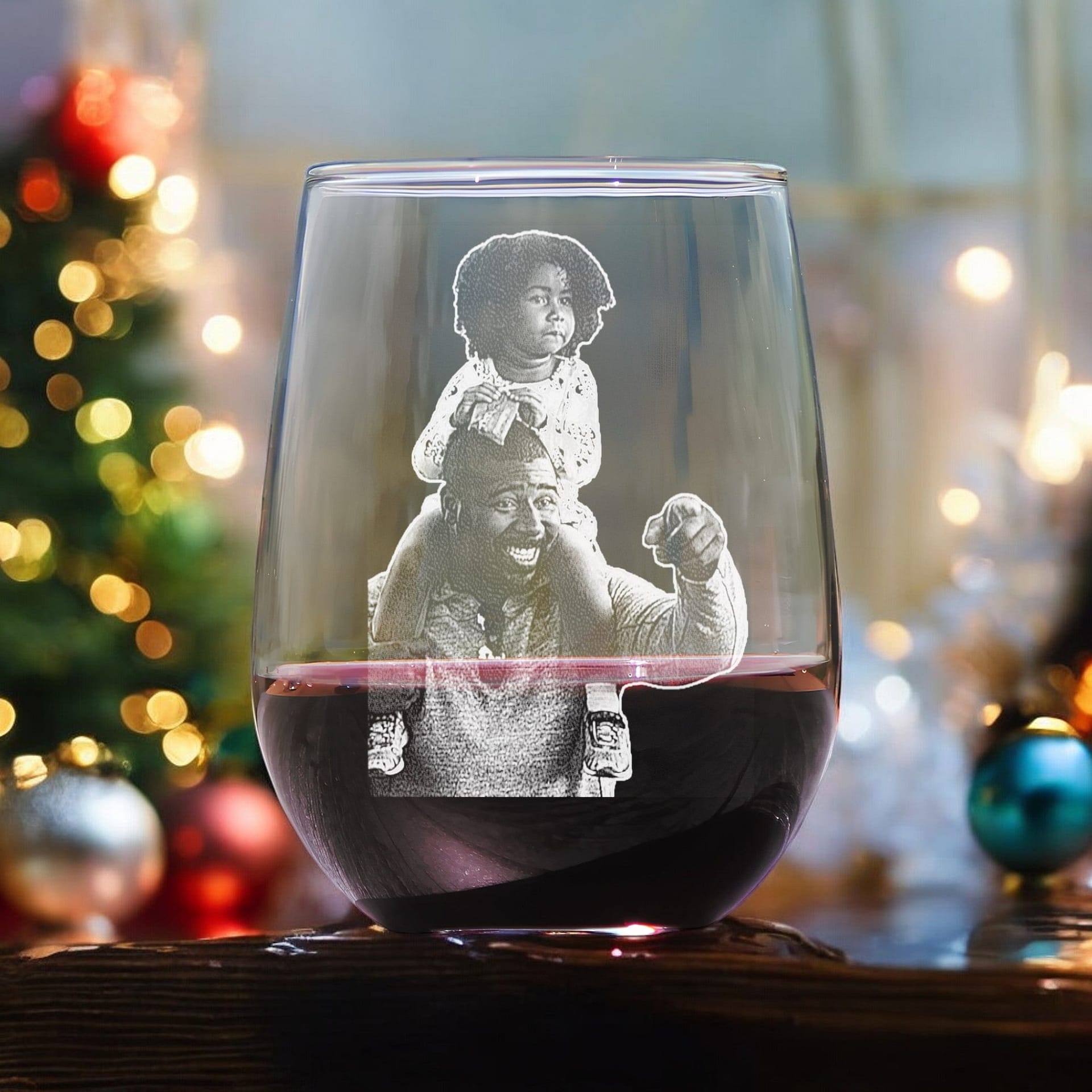 The First Father's Christmas Gift from My Store is a personalized stemless wine glass that showcases a black-and-white photo of a smiling adult holding a child on their shoulders, transforming this festive scene with a decorated Christmas tree and lights into an ideal custom gift for new dads.