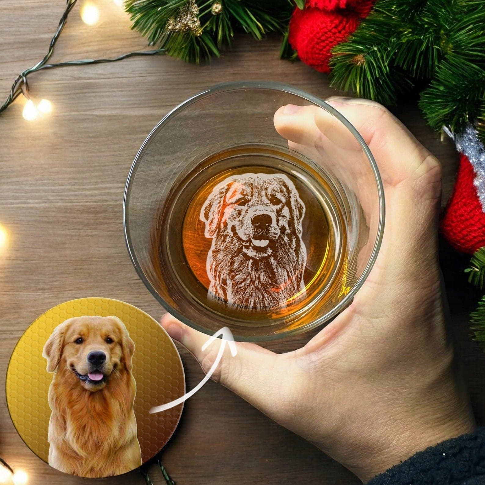 A hand holds the Christmas Custom Dog Photo Engraved Whiskey Glass, a personalized gift from My Store featuring an image of a Golden Retriever etched at the bottom. Beside this unique glass is a circular photo of the same dog. The wooden table is beautifully decorated with festive string lights.