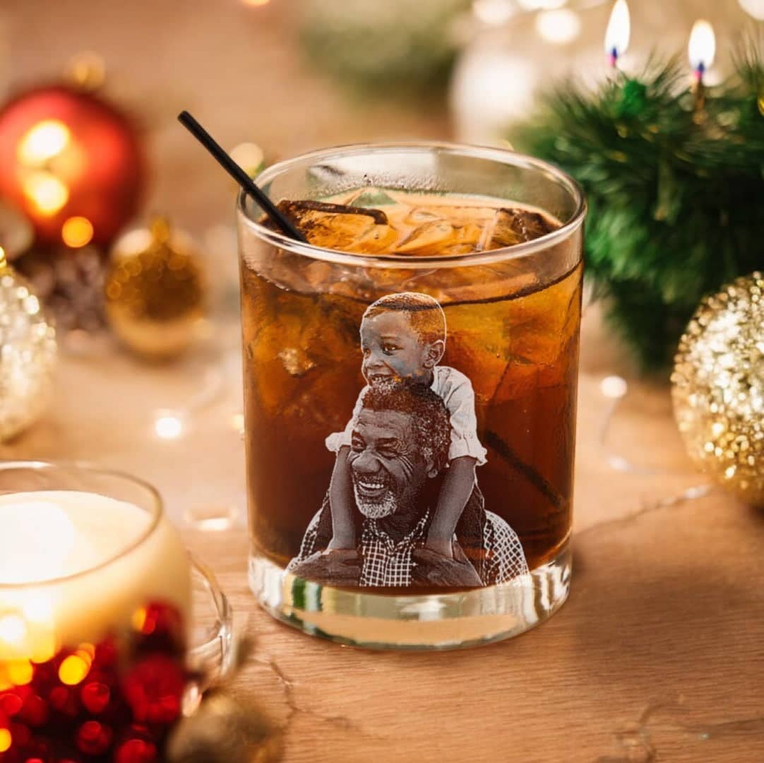 A festive scene showcases the "First Christmas as Grandpa" personalized 11oz whiskey glass from My Store, filled with a dark beverage, ice, and a black straw on a decorated table. This special glass features an illustration of a smiling man carrying a child on his shoulders to commemorate his first Christmas as grandpa. The setting is enhanced by lit candles and holiday ornaments surrounding the glass.