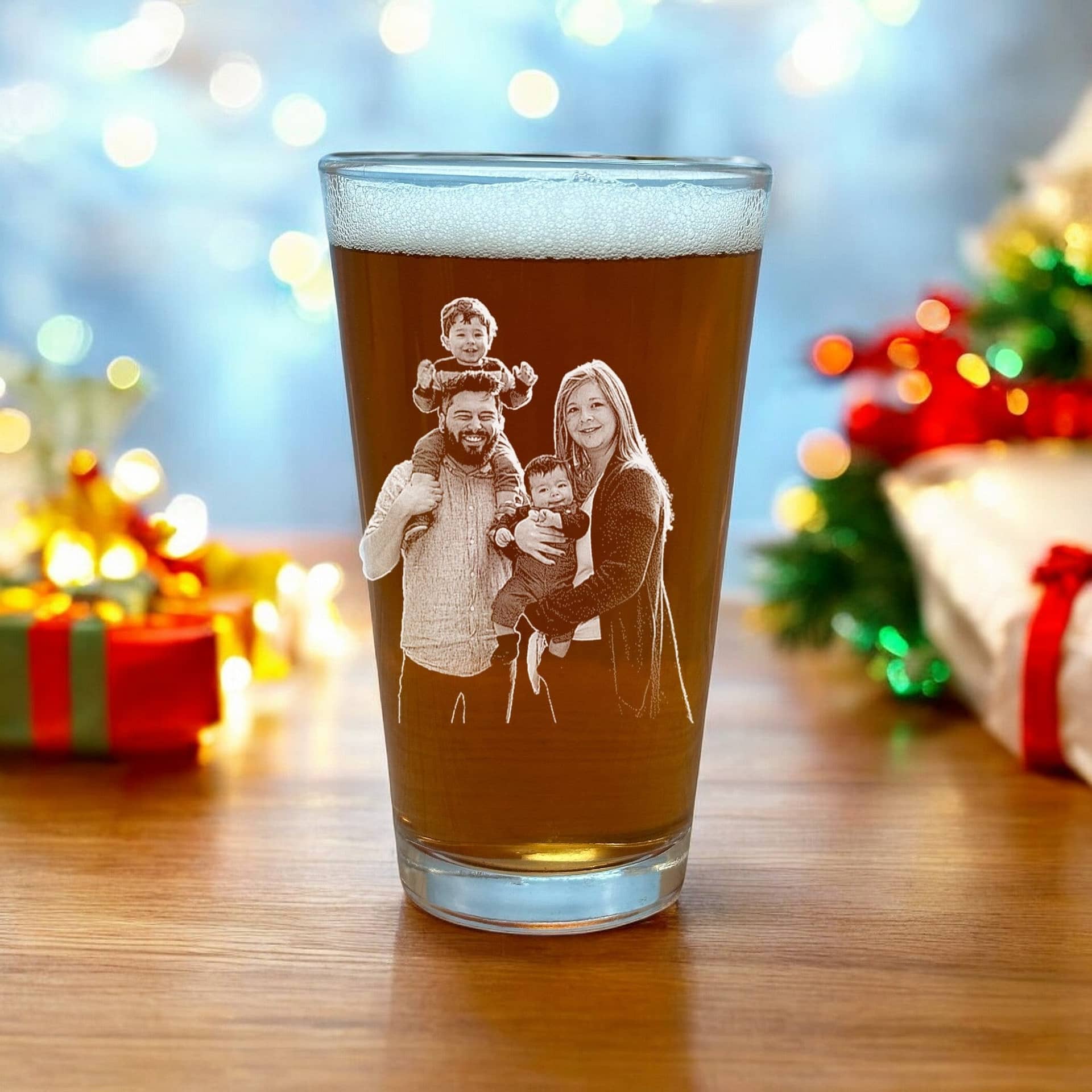 A 16oz beer pint glass from My Store's "First Christmas as Parents" collection, personalized and filled with a brown beverage, proudly displays a family photo of two adults and two children celebrating their first festive season together as parents. Behind the personal memento are softly blurred holiday decorations and gift boxes, enhancing the warmth of this cherished keepsake.